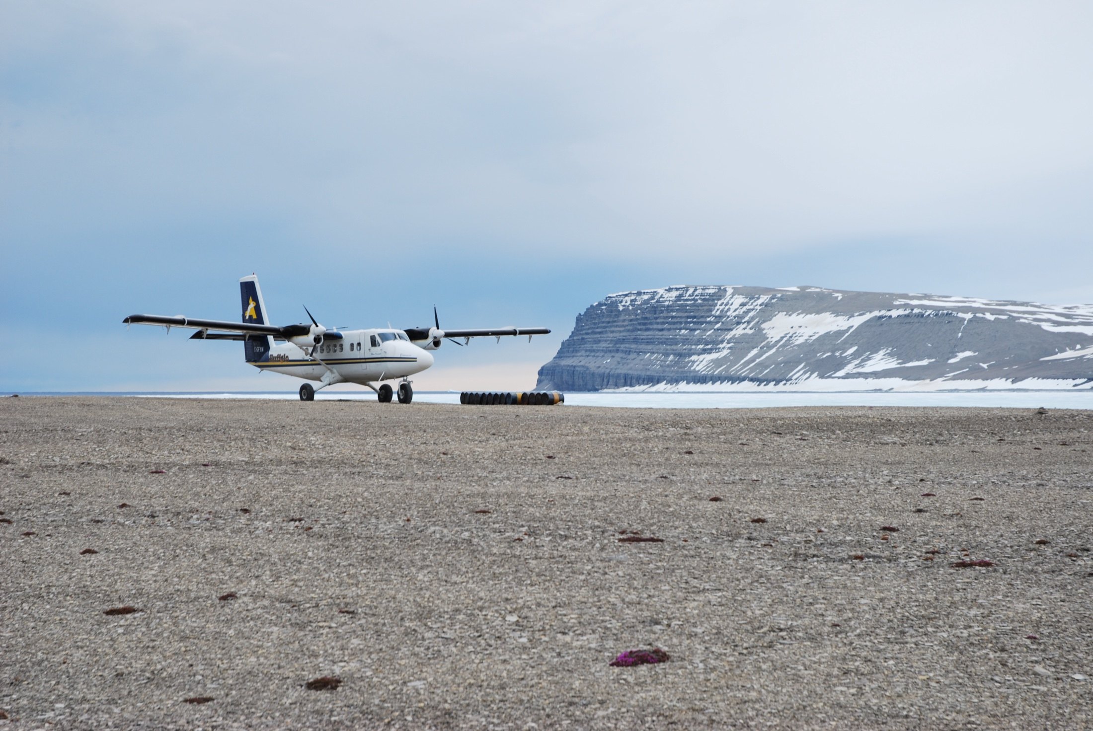 Parked on a gravel beach strip with fuel drums, sea ice and mesa cliffs beyond