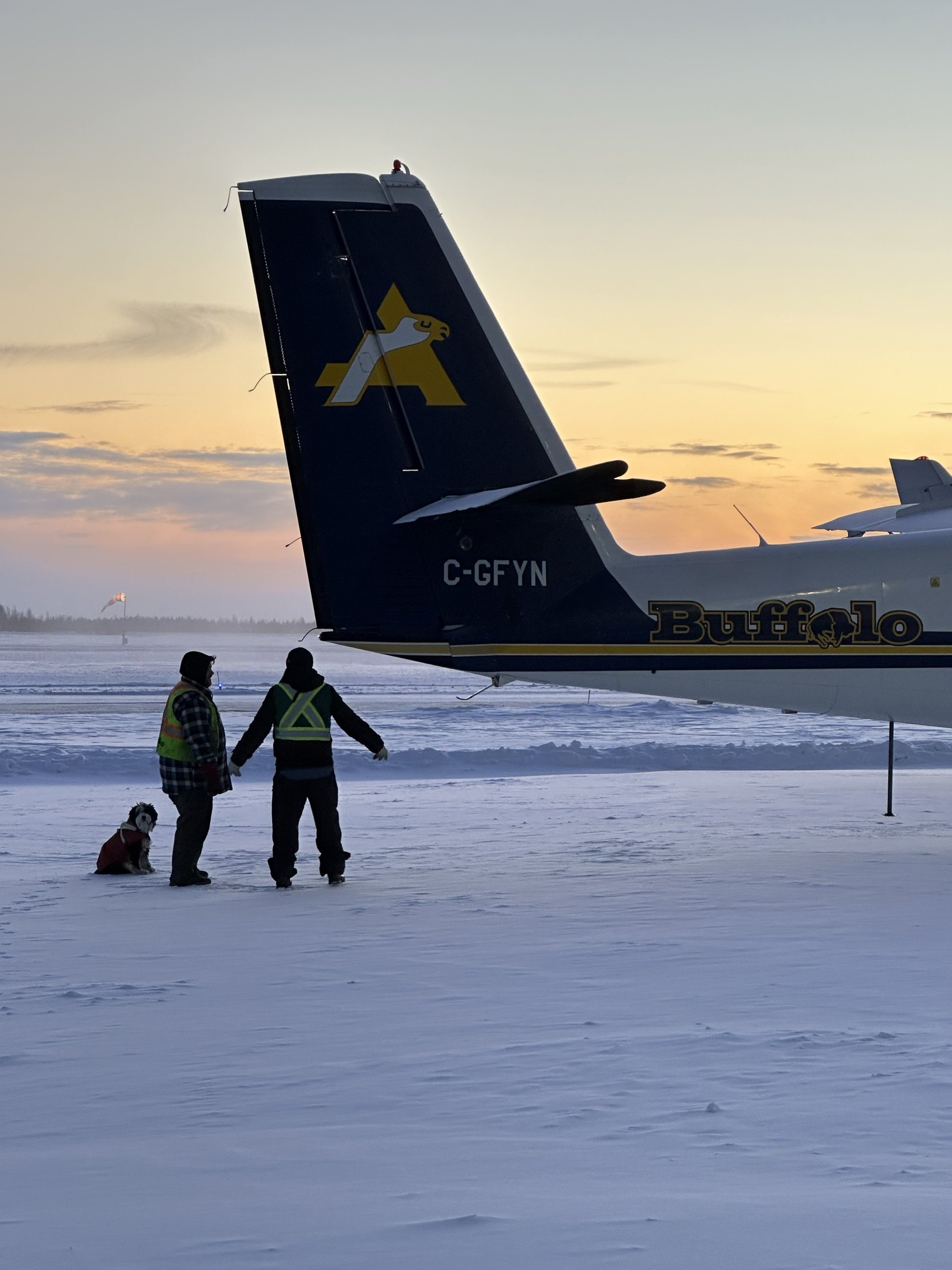 C-GFYN’s tail at winter sunset, two crew and a dog on the snow